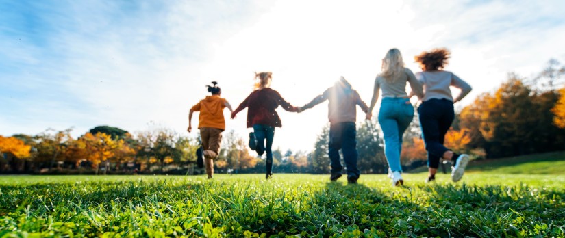 5 Menschen rennen Hand in Hand über eine Wiese auf die Sonne zu.
