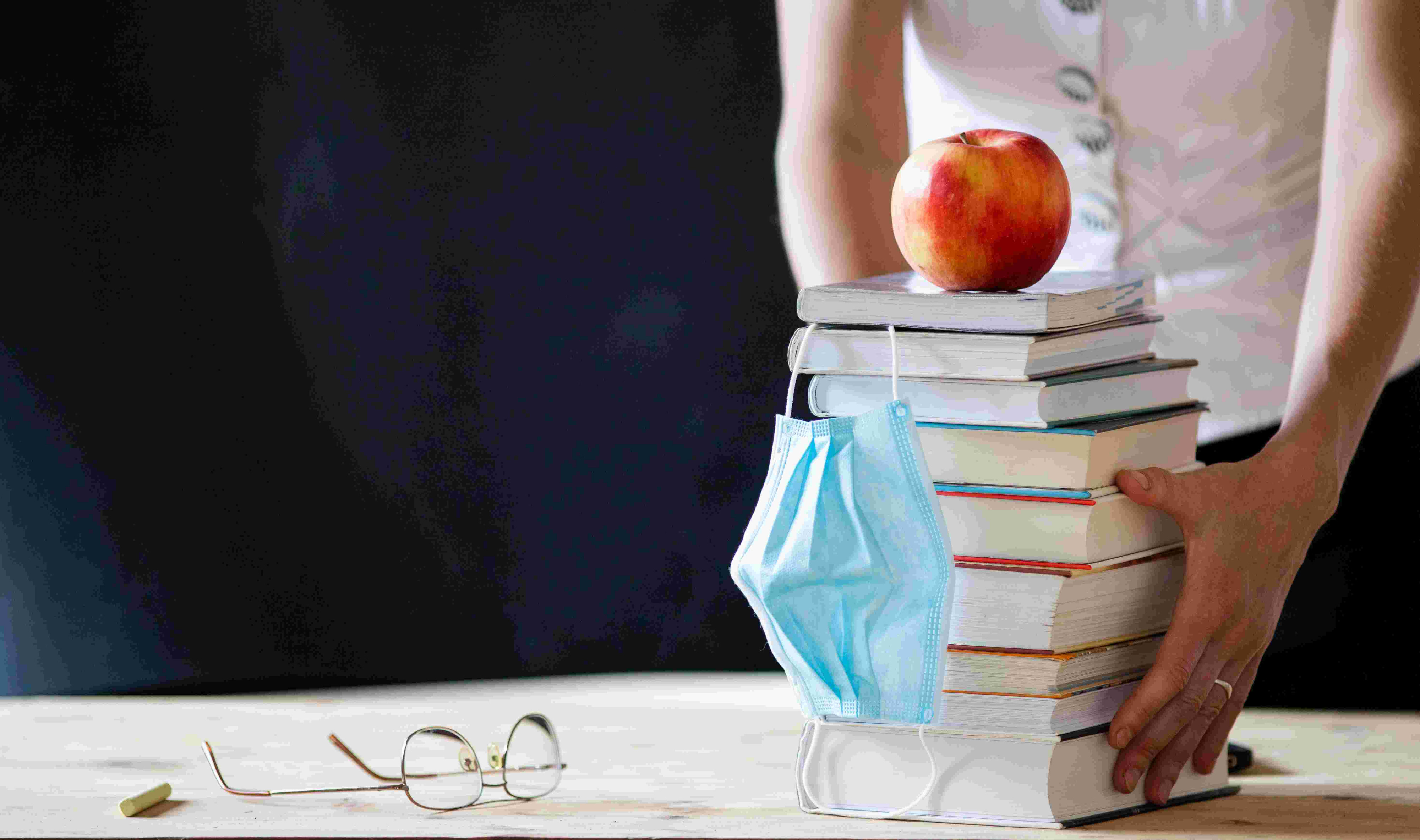 AUf einem Tisch liegen ein Bücherstapel mit Atemschutzmaske und einem Apfel, außerdem Papiere und eine Brille. Eine Person legt die Hände an den Bücherstapel
