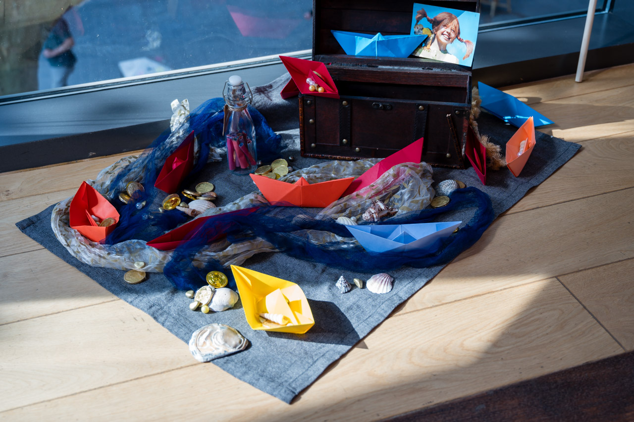 Tuch liegt am Boden mit maritimer Deko, wie Muscheln, Perlen, Goldthalern, Papierschiffen.
