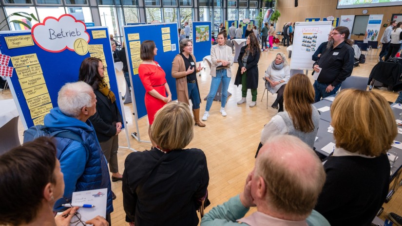 Menschen stehen um eine Pinnwand in einem großen Raum und hören einer Sprecherin zu 