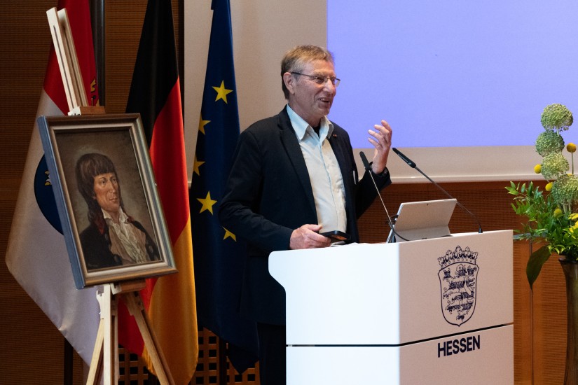 Der Laudator steht an einem Rednerpult. Neben ihm steht das Gemälde von Bernhard Christoph Faust auf einer Staffelei