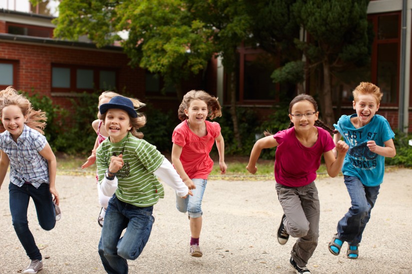 Sechs Kinder im Grundschulalter rennen mit Elan und Begeisterung auf einem Schulhof auf die Kamera zu. Im Hintergrund ist der Schulhof mit Bäumen und einem Gebäude zu sehen.