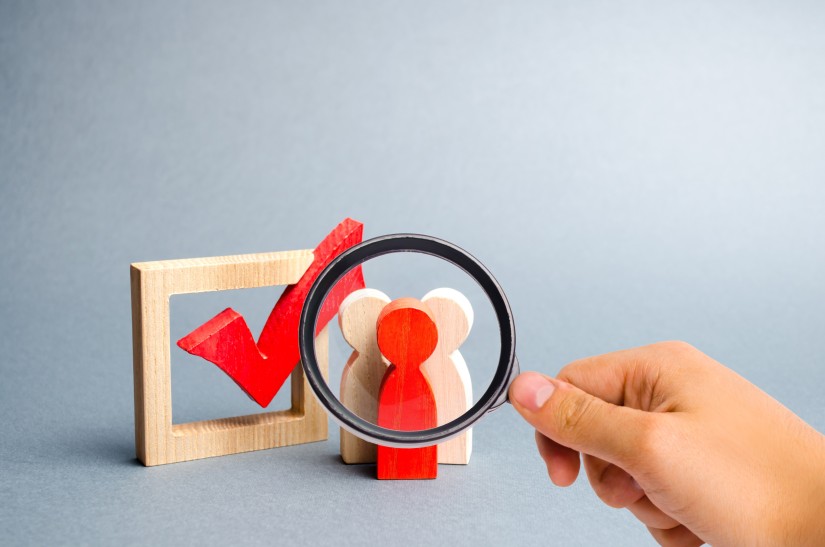 3 Holzfiguren vor einer Checkbox mit einer Lupe betrachtet